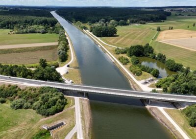 Eine moderne Brücke überspannt einen breiten Kanal inmitten von Feldern und Bäumen.