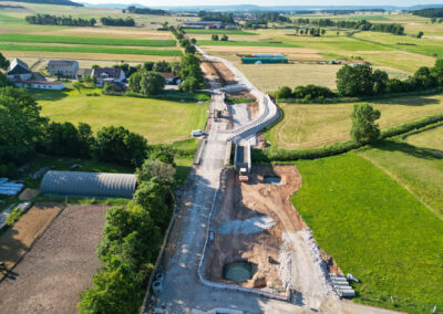 Luftaufnahme: ländlicher Straßenbau mit Maschinen, Feldern, Gebäuden, sonniger Tag.