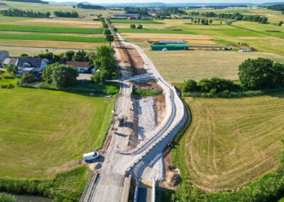 Luftaufnahme einer ländlichen Straßenbaustelle mit Maschinen, Feldern und Bäumen.