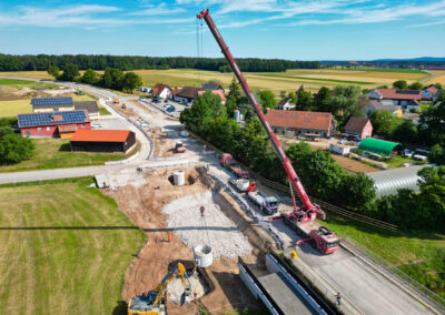 Ein Kran hebt Materialien; Fahrzeuge arbeiten auf der Straße. Häuser, Felder im Hintergrund.